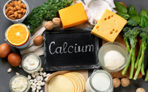 A chalkboard placard with the word "Calcium" written on it, with different food items like different kinds of cheese, and different leafy greens like kale, broccoli, as well as fruit like oranges, as well as nuts and seeds like almonds and walnuts, as well as dairies and milks, and meats, for a calcium rich diet for bone health.