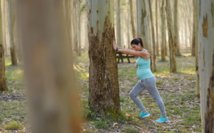 An outdoor scene of a pregnant person in baby blue tank top and gray slacks in a wooded area leaning with their arms out leaning against a tree and head slightly down.