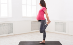 A pregnant person in a pink shirt and yoga pants standing on one leg, holding their foot behind their hip, in a bright room.