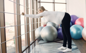 A pregnant person in black tights and a white long sleeved shirt leaning forward in a 90 degree angle, holding onto a window railing in front of exercise equipment.