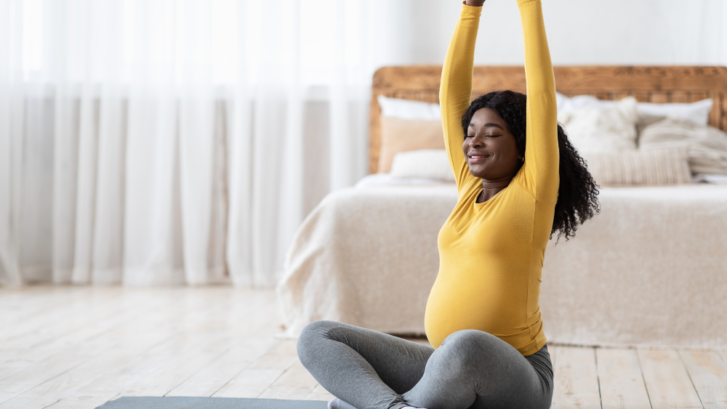 A dark skinned pregnant person in a bright yellow long sleeved shirt is happily stretching their arms up above their head while they sit cross legged in a well lit room on a yoga mat in what appears to be a bedroom.