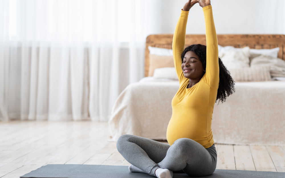 A dark skinned pregnant person in a bright yellow long sleeved shirt is happily stretching their arms up above their head while they sit cross legged in a well lit room on a yoga mat in what appears to be a bedroom.