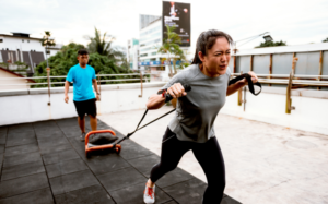 An Asian person straining while pulling a heavy object behind them, while another person behind them monitors their progress, as they practice their strength training.