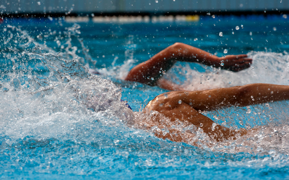 Two tanned people swimming in blue water, visible only from the waist up, their bodies splashing, using correct form to prevent swimming injuries