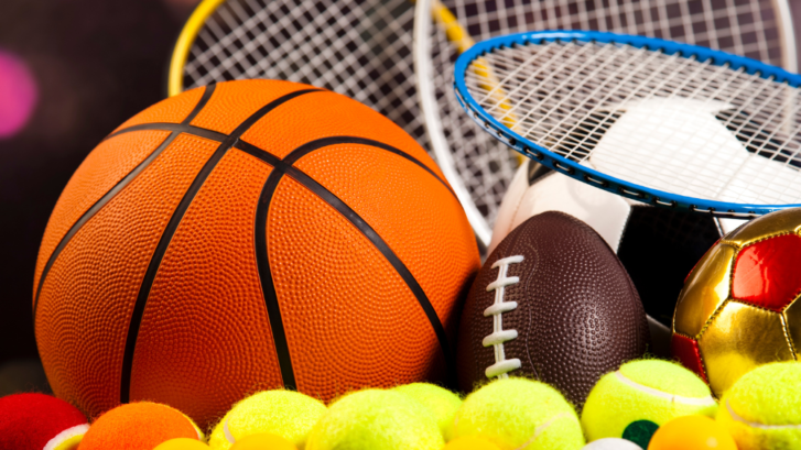 A close up image of a basketball, a football, tennis rackets, tennis balls, and a soccer ball, representing the world of sports