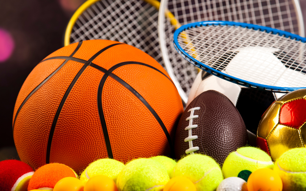 A close up image of a basketball, a football, tennis rackets, tennis balls, and a soccer ball, representing the world of sports