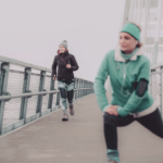 Picture of two people in workout gear on a pier, on a cold gray day. One person is wearing black workout leggings, a thick jacket, and a beanie, and is jogging in the background to the left. The other person is wearing a teal headband, a thick jacket, and leggings, and is stretching their knee forward.