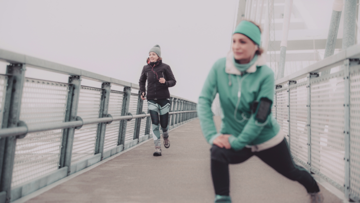 Picture of two people in workout gear on a pier, on a cold gray day. One person is wearing black workout leggings, a thick jacket, and a beanie, and is jogging in the background to the left. The other person is wearing a teal headband, a thick jacket, and leggings, and is stretching their knee forward.