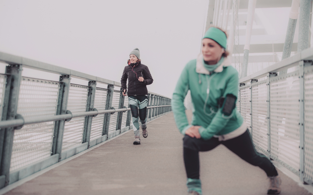 Picture of two people in workout gear on a pier, on a cold gray day. One person is wearing black workout leggings, a thick jacket, and a beanie, and is jogging in the background to the left. The other person is wearing a teal headband, a thick jacket, and leggings, and is stretching their knee forward.