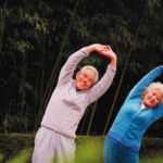 Photo of two older people, one in gray comfortable clothing, and one in blue, stretching with their arms above their heads, leaning to the side, in front a leafy green background.