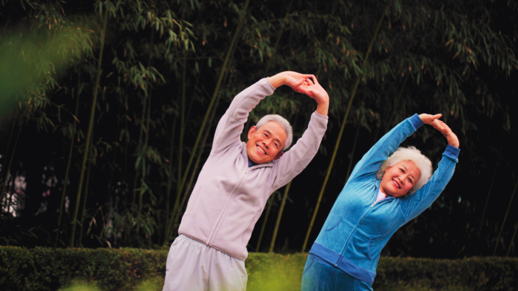 Photo of two older people, one in gray comfortable clothing, and one in blue, stretching with their arms above their heads, leaning to the side, in front a leafy green background.