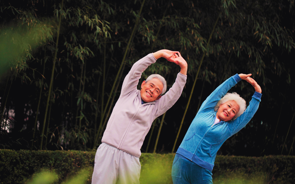 Photo of two older people, one in gray comfortable clothing, and one in blue, stretching with their arms above their heads, leaning to the side, in front a leafy green background.